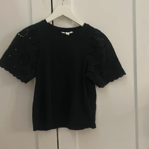 Svart blus med broderade ärmar XS - Svart t-shirt i storlek XS med korta, broderade ärmar och rund hals. Ärmarna har ett snyggt hålmönster och vågig kant som ger en cool touch. Perfekt för dig som gillar detaljer och vill ha något som sticker ut lite extra.