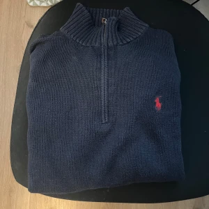  Polo Ralph Lauren zip - Snygg marinblå stickad polotröja från Polo Ralph Lauren med half zip och röd broderad logga på bröstet. Tröjan har ribbad krage och muddar, långärmad och perfekt för kyliga dagar