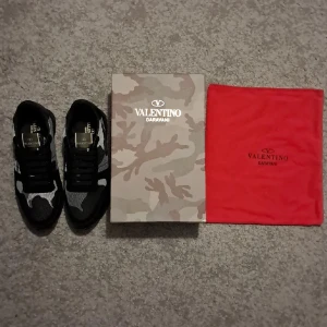 Valentino Garavani Rockrunner sneakers - Svarta och grå Valentino Garavani Rockrunner sneakers med camouflagemönster. Snygg mesh och mocka på ovandelen, platt sula med mönstrad undersida och snörning framtill. Ikonisk logga på plösen i metallic. Kommer med originalkartong och röd dustbag.