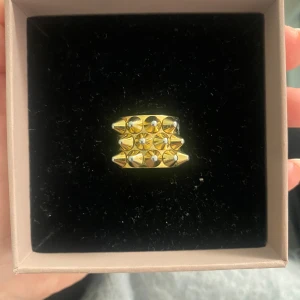 Bred guldfärgad ring med nitar - Cool ring i guldfärg med flera små konformade nitar i ett rutmönster. Säljer pga att jag bytt till silversmycken. Ringen saknar en av nitarna men det är inget som märks.(Därav priset). 
