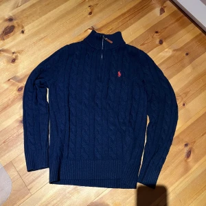 Marinblå stickad tröja Polo Ralph Lauren - Snygg marinblå stickad tröja från Polo Ralph Lauren med klassisk kabelstickning och röd logobrodyr på bröstet. Tröjan har half zip, ribbad krage och muddar samt långärmad passform. Perfekt för dig som gillar stilrena och tidlösa plagg.