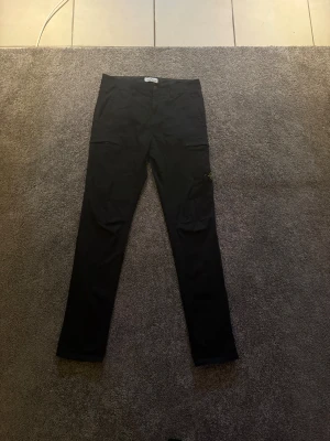 Svarta cargopants från Stone Island - Säljer ett par svarta cargopants från Stone Island med klassisk patch på benet. Byxorna har raka ben, flera fickor inklusive en med dragkedja och är gjorda i bomull. Perfekt för dig som gillar streetwear och vill ha en clean look med snygga detaljer. Dm för mer bilder och skriv frågor.