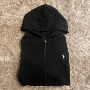 Svart hoodie från Polo Ralph Lauren - Svart hoodie från Polo Ralph Lauren med dragkedja och huva. Klassisk design med vit broderad logga på bröstet. Tröjan har väldigt bra skick. Men snörerna saknas
