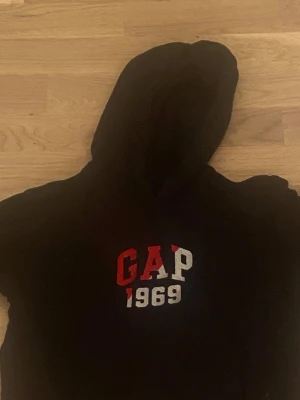 Svart GAP hoodie med tryck XL - Svart hoodie från GAP i storlek XL med klassiskt GAP 1969-tryck i rött och vitt på bröstet. Tröjan har huva och långa ärmar, perfekt för chill dagar. Materialet är mjukt och känns som bomull, vilket gör den skön att bära.