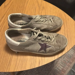 Golden Goose Superstar sneakers grå - Sunkiga Golden Goose Superstar sneakers i grå mocka med vit häl och lila stjärndetalj på sidan. Klassisk låg modell med snörning och platt sula. Märket syns på hälen och innersula 