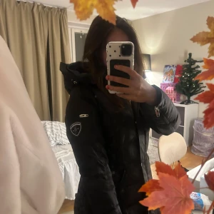 Svart dunjacka med huva från Rockandblue - Säljer en svart dunjacka från Rockandblue med snygga quiltade detaljer och huva. Jackan har silverfärgade dragkedjor, knappar och ett coolt märke på ärmen. Lång modell med flera fickor och justerbar huva, perfekt för kalla dagar.