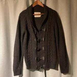 Svart stickad kofta från Le Costard - Säljer en snygg svart stickad kofta från Le Costard med kabelstickat mönster och knäppning framtill. Koftan har långa ärmar och ribbade muddar vid ärmslut och nederkant. Perfekt för kyliga dagar!