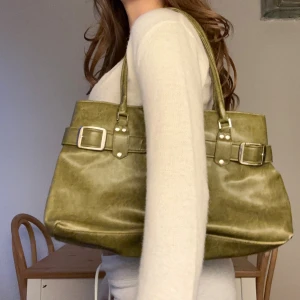 Olivgrön axelväska med spännen - Snygg olivgrön axelväska i skinnimitation med två rejäla handtag och coola silverfärgade spännen framtill. Väskan har en stilren, fyrkantig form och detaljerade sömmar som ger den en modern vibe. Perfekt storlek för att få plats med allt du behöver.