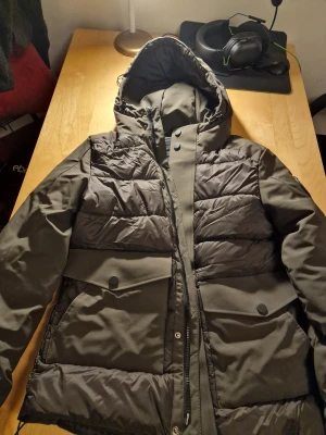 Mörkgrön pufferjacka S - Snygg mörkgrön pufferjacka från Smog i storlek XS men som kan även passa för S. Jackan har huva, stora fickor med knappar och coolt Outdoor Explorer-märke på ärmen. Materialet är polyester och syntet, med quiltade partier och dragkedja framtill. Perfekt för kalla dagar och chill stil.