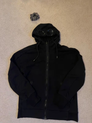 Svart hoodiejacka C.P. Company med goggles - Svart hoodiejacka från C.P. Company med ikoniska goggles i huvan. Jackan har dragkedja framtill, snörning vid huvan och två fickor med dragkedja. Materialet är mjuk bomull och passformen är normal. Perfekt för dig som vill sticka ut med streetwear-vibe. Äkta med qr kod som scannar