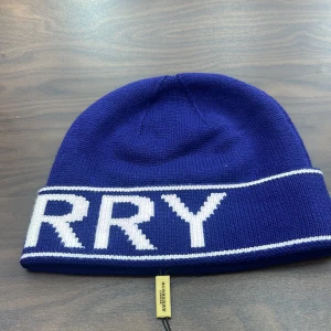 Blå stickad Burberry mössa - Snygg marinblå stickad mössa från Burberry med vit logotyp runt om. Klassisk form med uppvikt kant och tydlig Burberry-tag. Perfekt för dig som vill ha en cool och lyxig accessoar med tydlig branding.