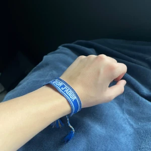 Blått Christian Dior armband - Snyggt blått armband från Christian Dior med texten 'J'Adior' broderad i vitt. Armbandet har flätade band och två blå tofsar som detalj. Perfekt accessoar för att lyfta din outfit med en lyxig touch och cool design.