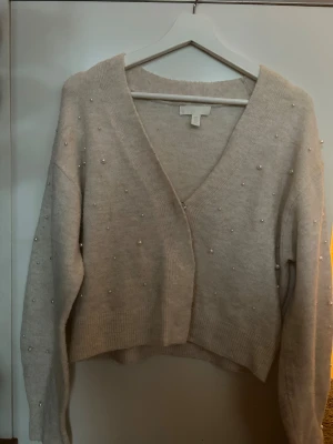 Beige kofta med pärlor från H&M - Superfin beige kofta från H&M med v-ringning och croppad passform. Tröjan är dekorerad med små pärlor över hela och har långa ärmar samt breda ribbade muddar. Perfekt för dig som gillar mjuka material och snygga detaljer.