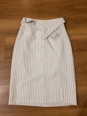 Vit randig långkjol från Zarina - Snygg vit långkjol från Zarina Collection med svarta vertikala ränder och ett matchande bälte i midjan. Kjolen har en slits framtill som ger extra edge och är tillverkad i ett mjukt polyestermaterial. Perfekt för dig som gillar stilrena och moderna plagg. EU-storlek 38 (står det 44)