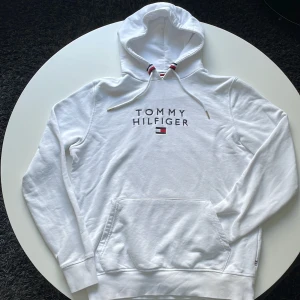 Vit hoodie Tommy Hilfiger strl M - Snygg vit hoodie från Tommy Hilfiger med broderad logga på bröstet och klassiska Hilfiger-detaljer i rött, vitt och blått vid snörningen. Tröjan har huva, känguruficka och långa ärmar. Perfekt för chill dagar och streetwear.