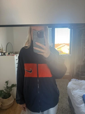 Nike vindjacka i mörkblå och röd - Nike vindjacka i mörkblått med en bred röd panel över bröstet och klassisk vit swoosh-logga. Jackan har dragkedja framtill, hög krage och långärmade ärmar. Perfekt för dig som gillar sportig och chill stil.
