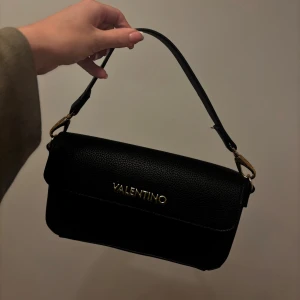 Svart axelväska från Valentino - Snygg svart axelväska från Valentino i stilren design med guldfärgade detaljer och präglat skinn. Väskan har ett praktiskt innerfack med dragkedja och magnetlås. Köptes på zalando 2023 