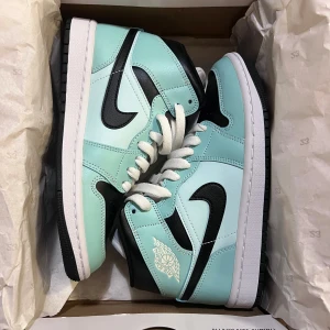 Nike Air Jordan 1 Mid Teal Tint - Nike Air Jordan 1 Mid i färgen teal tint med svarta och vita detaljer. Skorna har klassisk snörning, svart Swoosh och perforerad tå. Ovandelen är i skinn och modellen har en vit mellansula med svart yttersula. Perfekt för dig som vill sticka ut med snygga sneakers.