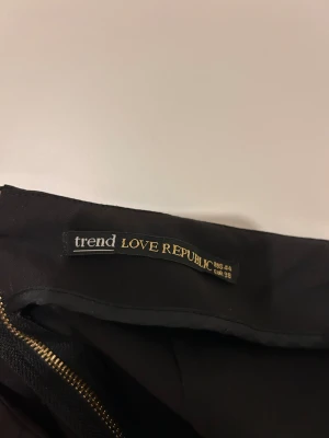 Svart omlottkjol från Love Republic - Svart omlottkjol från Love Republic med elegant drapering framtill och en liten metallknapp som detalj. Kjolen har en stilren, rak passform och är knälång. Perfekt för dig som gillar minimalistisk och chic stil.