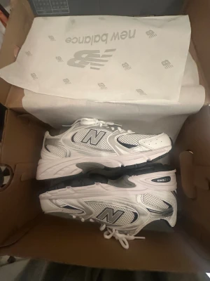 New Balance 530 vita sneakers - Säljer ett par New Balance 530 sneakers i vitt med silver- och grå detaljer. Skorna har meshpaneler för extra andningsförmåga, klassisk snörning och en chunky sula i svart och vitt. Perfekta för dig som gillar retrovibbar och sportig stil. Jag själv använder aldrig dem de ligger bara i garderoben 