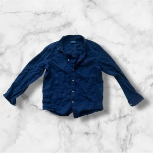 Marinblå skjorta från Polo Ralph Lauren - Snygg marinblå skjorta från Polo Ralph Lauren i storlek 18-20 men passar jättebra S! Skjortan har klassisk krage, knappar framtill och lång ärm. Tillverkad i mjuk bomull och har den ikoniska röda Polo-loggan broderad på bröstet. Perfekt för dig som gillar stilrena och tidlösa plagg.