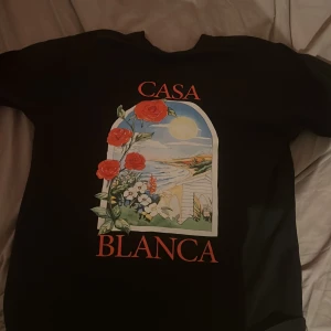 Svart t-shirt från Casablanca M - Svart t-shirt från Casablanca med färgglad tryckt grafik framtill som visar rosor och strandmotiv i ett fönster. T-shirten har rund hals och korta ärmar. Snyggt röd texttryck 'CASA BLANCA' på bröstet. Perfekt för dig som gillar unika prints.
