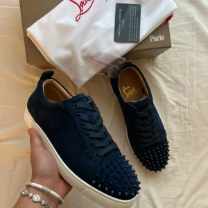Mörkblå Christian Louboutin sneakers - Säljer ett par mörkblå sneakers från Christian Louboutin med coola nitar på tån och klassisk röd sula. Skorna har snörning, rund tå och är tillverkade i mocka med vita platta sulor. Perfekt för dig som vill sticka ut med exklusiv stil. Helt nya aldrig använda.Skriv om ni har några frågor eller funderingar 😊