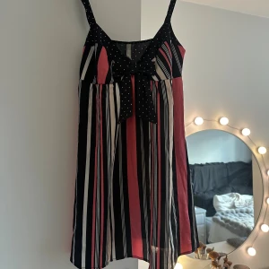 Kort söt klänning - Snygg kort klänning med smala axelband, svart överdel med vita prickar och en söt rosett framtill. Kjoldelen har vertikala ränder i svart, rosa och vitt. Lätt och luftig modell, perfekt för varma dagar.