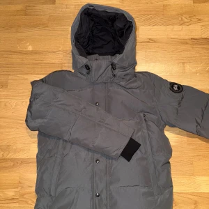 Canada Goose Jacket Grey - Säljer en grå dunjacka från Canada Goose med stor huva, svarta detaljer och patch på ärmen. Jackan har dragkedja, knappar och medföljande päls till huvan. Perfekt för kalla vinterdagar och har en clean, modern look.
