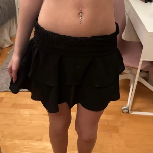 Svart volangkjol med smock - Supersöt svart kortkjol med bred smock i midjan och dubbla volanger. Kjolen har en flowy känsla och sitter bekvämt tack vare den elastiska midjan. Perfekt att styla med crop tops eller hoodies. Materialet känns lätt och luftigt.