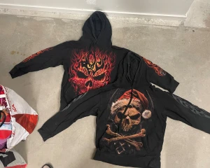 Svarta hoodies med dödskalle från Spiral M/S - Två svarta hoodies från Spiral med stora tryck av dödskallar. Ena har eldflammor och dödskallemotiv, den andra har dödskalle med tomteluva och korslagda ben. Båda har huva och tryck på ärmarna. Perfekt för dig som gillar alternativ stil. Kan även säljas styckvis 