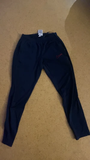 Blå Nike träningsbyxor M - Snygga blå träningsbyxor från Nike i storlek M. Byxorna har ett rött Nike Swoosh broderat på benet och är gjorda i ett lätt och stretchigt syntetmaterial. De har smal passform och elastisk midja med snörning för extra komfort.