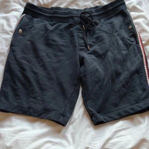 Svarta shorts från Moncler XL - Svarta shorts från Moncler i mjuk bomull med snörning i midjan och silverfärgade knappar med logga. Sidorna har randiga detaljer i rött och vitt. Perfekta för chill dagar och sportiga outfits.