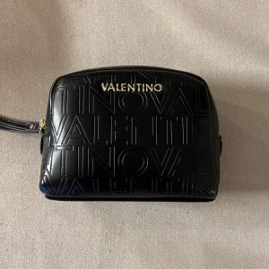 Valentino väska svart - Snygg svart väska från Valentino med präglad logotyp över hela väskan och guldfärgad metalltext framtill. Väskan har en rektangulär form, dragkedja upptill och justerbar axelrem. Perfekt storlek för det viktigaste och riktigt stilren design. I princip aldrig använd. Plast kvar på dragkedjan inuti. Först till kvarn! 