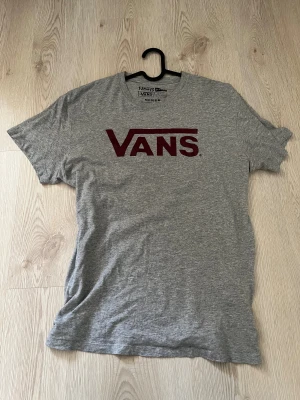 Grå Vans t-shirt med röd logga - Snygg grå t-shirt från Vans i storlek medium med klassisk röd Vans-logga på bröstet. T-shirten har rund hals och korta ärmar. Materialet är mjuk bomull med lite polyester, perfekt för en chill och avslappnad stil.