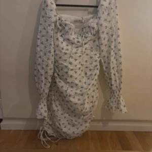 Vit blommig långklänning från Adoore - Superfin långklänning från Adoore i vit med blått blommigt mönster. Klänningen har långa puffärmar, snörning framtill och volang vid ärmslut. Perfekt för sommarens alla tillfällen, med romantisk vibe och snygg passform.