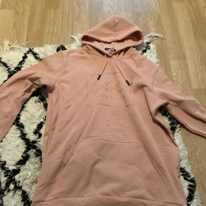 Rosa hoodie från True North - Mysig rosa hoodie från True North med broderad logga och bergmotiv på bröstet. Tröjan har huva med snörning och en stor magficka. Perfekt för chill dagar och passar till allt. Tillverkad i mjuk bomullsmix för extra komfort. 158-164  kan sänka pris