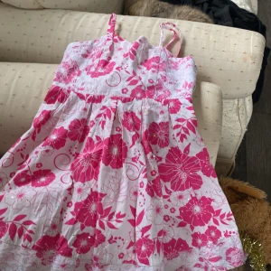 Rosa blommig kortklänning med spets - Supersöt kortklänning med rosa och vita blommor, tunna axelband och spetsdetaljer upptill och längst ner. Klänningen har glittriga små stenar och är perfekt för varma dagar. Materialet känns lätt och luftigt, och passformen är normal.