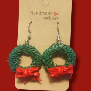 Juliga örhängen med krans och rosett - Unika handgjorda örhängen formade som gröna julkransar med detaljerade röda rosetter. Perfekt för dig som vill sticka ut med en festlig och kreativ look under julen. Örhängena har silvriga krokar och är tillverkade i polymerlera.