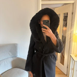 Svart kappa med stor pälshuva - Svart kappa med stor fluffig pälshuva och knytbälte i midjan. Dubbelknäppt framtill och raka, långärmade ärmar. Snygg och stilren från HM