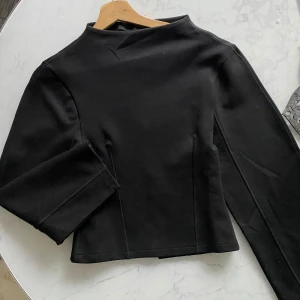 Svart tröja med funnel neck XS - Minimalistisk svart figursydd tröja från gina i storlek XS. Toppen har en hög funnel neck och långa ärmar, sydd i ett mjukt och stretchigt material som ger en clean och stilren look. Perfekt att styla med jeans eller kjol för en modern vibe.