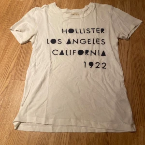 Vit t-shirt från Hollister XS - Vit t-shirt från Hollister i storlek XS med svart tryck på framsidan där det står 'Hollister Los Angeles California 1922'. Klassisk rund halsringning och korta ärmar. 