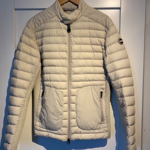 Beige pufferjacka från Colmar Originals - Snygg beige pufferjacka från Colmar Originals med snygg design. Jackan har två stora fickor med dragkedja framtill och en diskret logotyp på ärmen. Perfekt för dig som vill ha en clean och stilren look under höst och vinter.