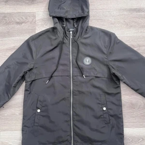 Svart vindjacka från Trapstar - Svart vindjacka från Trapstar med huva och dragkedja framtill. Jackan har logotyp på bröstet och broderad Trapstar-text på ryggen. Två fickor med knappstängning och justerbar snörning vid huvan. Materialet är lätt och syntetiskt, perfekt för streetwear.