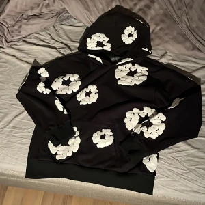 Svart hoodie med vita blommor Denim Tears - Svart hoodie från Denim Tears med stora vita blommor tryckta över hela tröjan. Tröjan har huva, långa ärmar och ribbade muddar. 