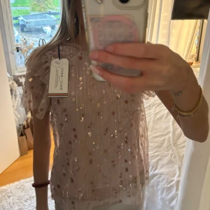 Glittrig rosa blus  - Säljer en superfin ljusrosa blus från Indiska med glittriga paljetter i guld och silver. Blusen har korta, volangiga ärmar och är tillverkad i ett tunt, transparent syntetmaterial. Perfekt för att sticka ut med extra shimmer och volym på axlarna. Defekter syns på bilden