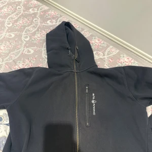 Svart hoodie från Sail Racing XL - Svart hoodie från Sail Racing i storlek XL med dragkedja hela vägen, bröstficka med dragkedja och broderad logga. Mjuk fleece på insidan och huva med snörning. Perfekt för kyliga dagar och riktigt skön att ha på sig.