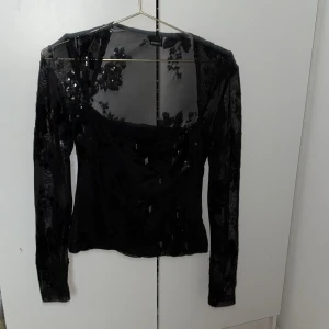 Svart mesh blus med paljetter - Säljer en svart mesh blus från Ginatricot med långärmade transparenta ärmar och blommigt paljettbroderi. Blusen har en croppad passform och fyrkantig urringning. Perfekt för dig som gillar detaljer och vill sticka ut med glittriga inslag.