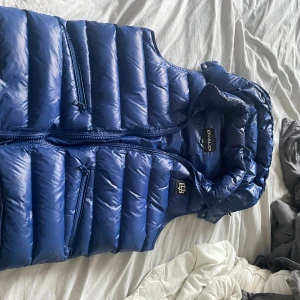 Bvaldi väst  - Snygg blå dunväst från BVALDI med quiltad design och huva. Västen har två dragkedjeförsedda fickor framtill och en cool logga på bröstet. Perfekt för lager-på-lager och streetstyle. Stängs med dragkedja hela vägen.