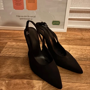 Svarta pumps med spetsig tå från Bershka - Säljer ett par svarta pumps från Bershka i storlek 41. Skorna har spetsig tå, öppen häl med rem och låg klack. Materialet är syntet och de har en stilren, elegant look som passar till många outfits.